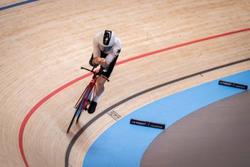 Ein weiß-schwarz gekleideter Radfahrer fährt auf einem roten Fahrrad auf einer Indoor-Radrennbahn und lehnt sich in eine Kurve, die mit blauen, schwarzen und roten Linien markiert ist.