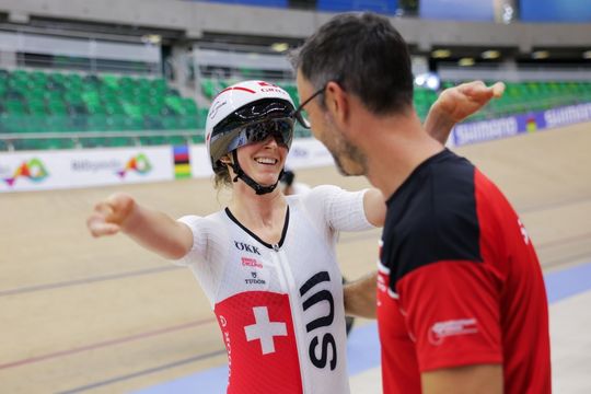 Ein lächelnder Radfahrer in Schweizer Teamkleidung und Helm umarmt einen Mann in einem rot-schwarzen Trikot in einer Radrennhalle mit leeren grünen Sitzen im Hintergrund.