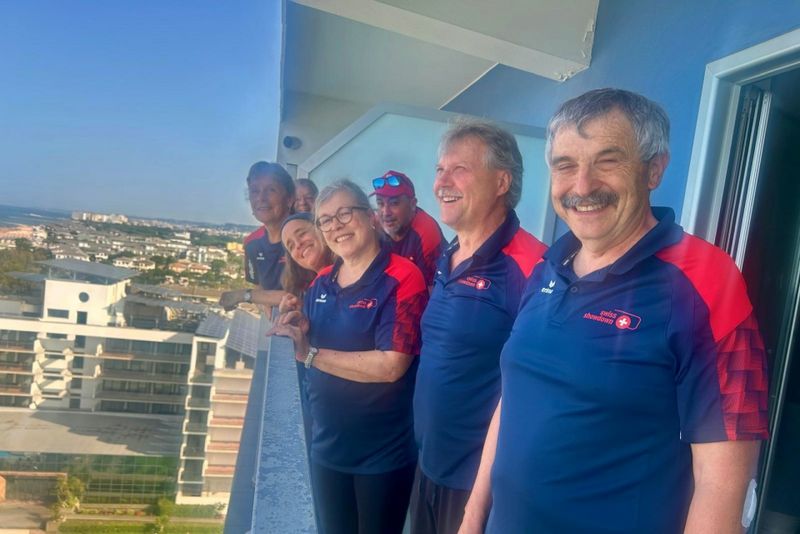 Eine Gruppe von sechs lächelnden Erwachsenen in passenden marineblauen und roten Hemden steht an einem sonnigen Tag auf einem hohen Balkon mit Blick auf eine Stadtlandschaft und die Küste.