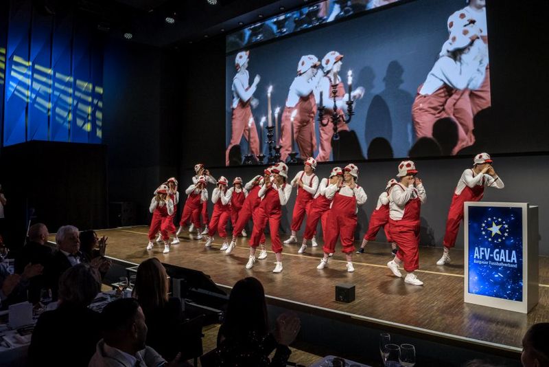 Eine Gruppe von Künstlern in passenden roten Overalls und Hüten tanzt auf der Bühne der AFV-Gala, während das Publikum zuschaut und ein großer Bildschirm im Hintergrund eine Nahaufnahme der Aufführung zeigt.