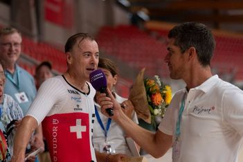 Ein männlicher Sportler in einem rot-weißen Schweizer Trikot wird von einem Reporter mit einem lila Mikrofon interviewt. Im Hintergrund sind Menschen und Blumen zu sehen, die von roten Stadionsitzen umgeben sind.