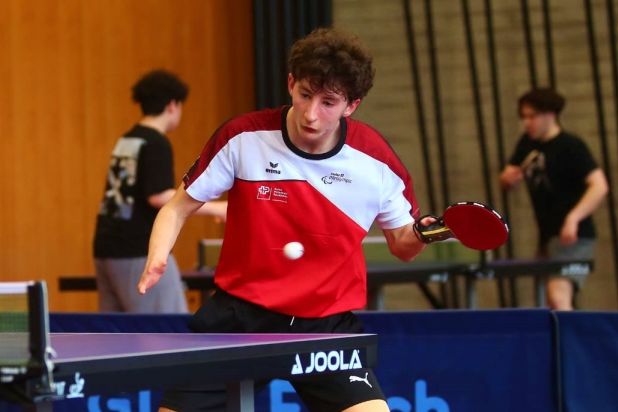 Ein junger männlicher Tischtennisspieler mit Beinprothese in Aktion, der ein rot-weißes Hemd und schwarze Shorts trägt und gerade einen Schlag abgibt. Zwei weitere Personen stehen im Hintergrund an einem anderen Tisch.