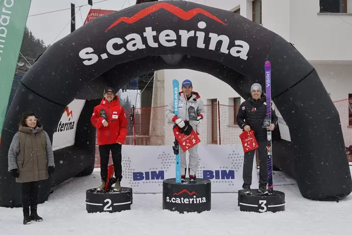 Drei Athleten stehen auf einem verschneiten Siegertreppchen unter einem schwarzen Bogen der Heiligen Caterina und halten Preise in der Hand. Der Athlet in der Mitte belegt den ersten Platz, flankiert von den Zweit- und Drittplatzierten. Es fällt Schnee; links steht eine Frau.