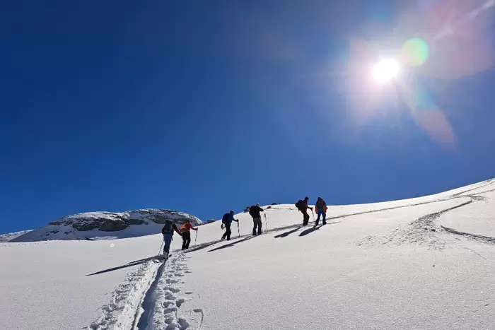 Eine Gruppe von Menschen wandert bei strahlend blauem Himmel einen verschneiten Berghang hinauf, während über ihnen die Sonne scheint und die Skispuren im Schnee sichtbar sind.
