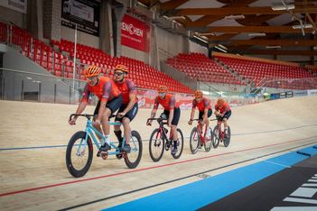 Fünf Radfahrer mit passenden orangefarbenen Helmen und roten Trikots fahren in einer Reihe auf einer hölzernen Indoor-Velodrombahn, im Hintergrund leere rote Stadionsitze.