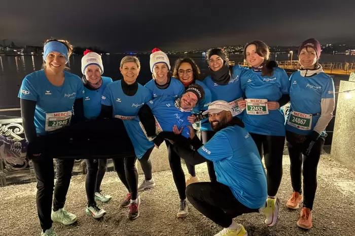 Eine Gruppe von elf lächelnden Läufern in passenden blauen Hemden posiert nachts zusammen und hält einen Teamkollegen waagerecht hoch. Sie tragen Startnummern und Wintermützen und stehen an einer Uferpromenade mit den Lichtern der Stadt im Hintergrund.