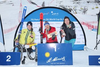 Drei Para-Alpin-Skisportlerinnen feiern auf einem Podium im verschneiten St. Moritz, in der Hand ihre Medaillen, Skier und Preise. Die Siegerin sitzt lächelnd in der Mitte des obersten Podestplatzes, die anderen stehen auf beiden Seiten. Hinter ihnen sind die Veranstaltungsbanner zu sehen.