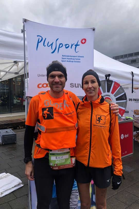 Zwei Läufer in orangefarbenen Hemden, eines davon mit einem "GUIDE"-Aufkleber und einer Startnummer, stehen lächelnd nebeneinander bei einer Sportveranstaltung. Im Hintergrund sind ein Plusport-Banner und verschiedene Sponsorenlogos zu sehen.