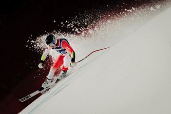 Ein Skifahrer in rot-weißem Anzug und Helm rast bei einem Wettkampf auf einer steilen, verschneiten Piste bergab, schneidet scharf und wirbelt eine Schneeschaufel auf.