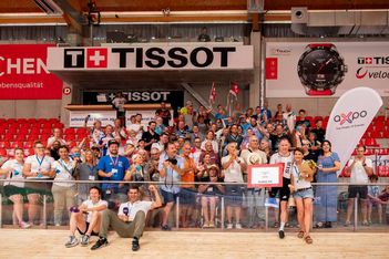 Eine grosse Gruppe von Menschen posiert und lächelt zusammen in einer Sporthalle und hält Transparente und Schilder. Die Firmenlogos von Tissot und Axpo sind über und hinter ihnen zu sehen, ebenso wie die rote Stadionbestuhlung.
