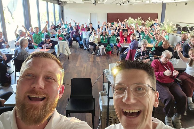 Zwei Männer im Vordergrund machen ein lächelndes Selfie mit einer großen, fröhlichen Gruppe von Menschen, die an Tischen hinter ihnen sitzen, viele heben ihre Hände und winken in einem hellen, modernen Raum.
