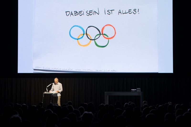 Eine Person steht auf einem Podium auf der Bühne und spricht zum Publikum. Dahinter ist eine große Leinwand mit den olympischen Ringen und dem deutschen Spruch „Dabeisein ist alles!“ zu sehen.