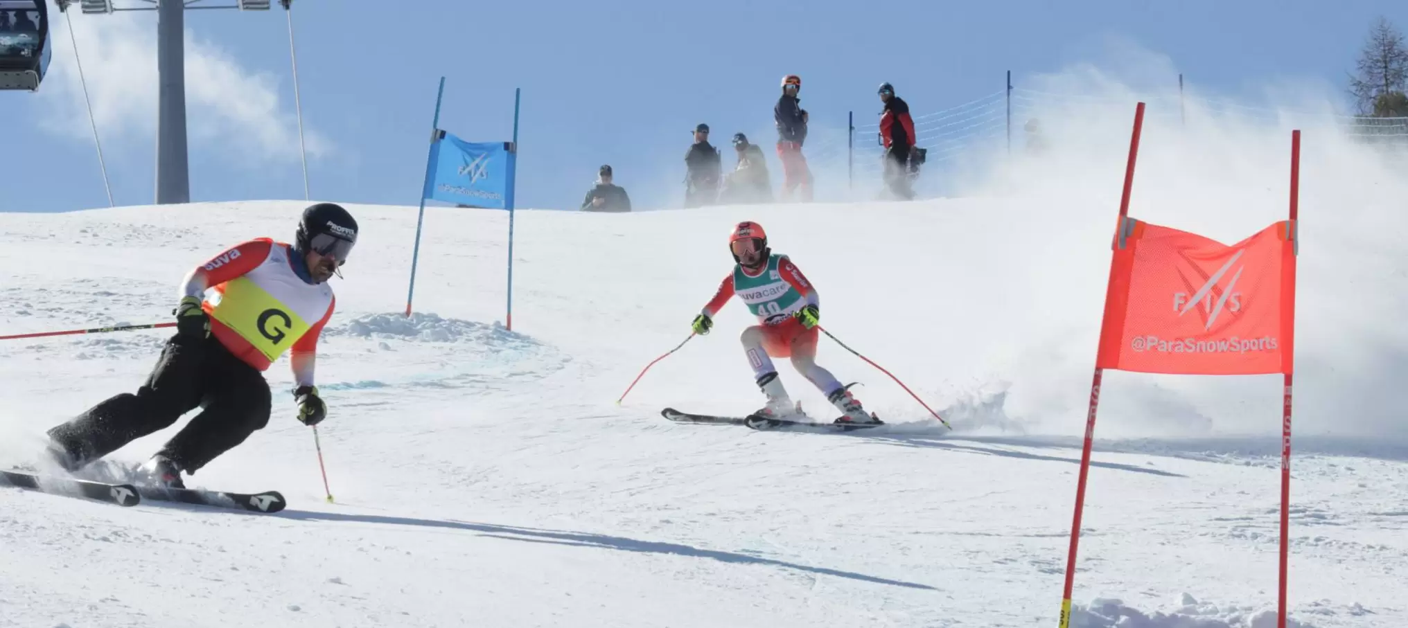 Zwei Skifahrer, die nummerierte Startnummern tragen, rasen einen verschneiten Hang durch Tore hinunter, mit blauem Himmel und Zuschauern im Hintergrund. Der führende Skifahrer trägt eine gelbe "G"-Staffel; beide stoßen bei ihren Kurven Schnee auf.