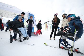 Eine Gruppe von Personen, darunter zwei Personen mit adaptiver Skiausrüstung, versammelt sich auf verschneitem Boden in der Nähe von Zelten und Fahnen. Einige stehen und andere sitzen, alle in Winterkleidung, in einem Skigebiet mit Bergen im Hintergrund.