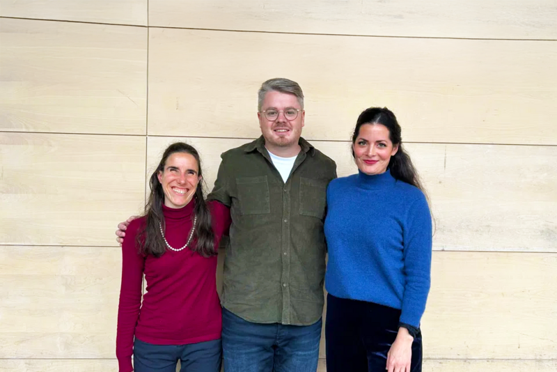 Drei Erwachsene stehen lächelnd vor einer hellen Holzwand. Die Person links trägt einen roten Rollkragenpullover, die Person in der Mitte eine Brille und ein grünes Hemd und die Person rechts einen blauen Pullover.