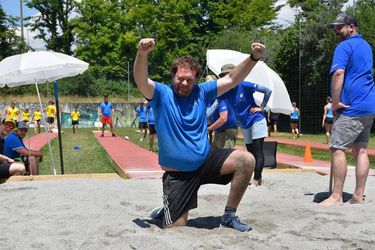 Ein Mann in blauem Hemd und Shorts kniet auf sandigem Boden und hebt feierlich die Arme. Menschen in blauen und gelben Hemden schauen aus dem Hintergrund auf einem sonnigen Sportplatz im Freien zu.