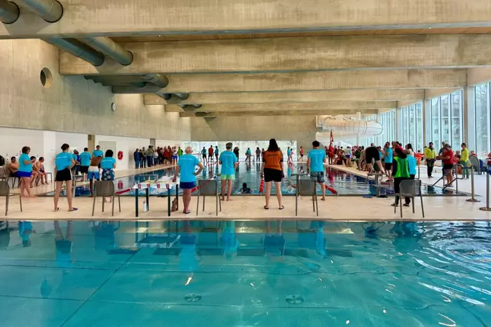 Ein Schwimmwettkampf in einem Hallenbad mit Schwimmern, Trainern und Zuschauern. Die Leute stehen am Beckenrand, während andere hinter Absperrungen zuschauen. Der Schwimmbereich hat große Fenster und Betonbalken über dem Kopf.