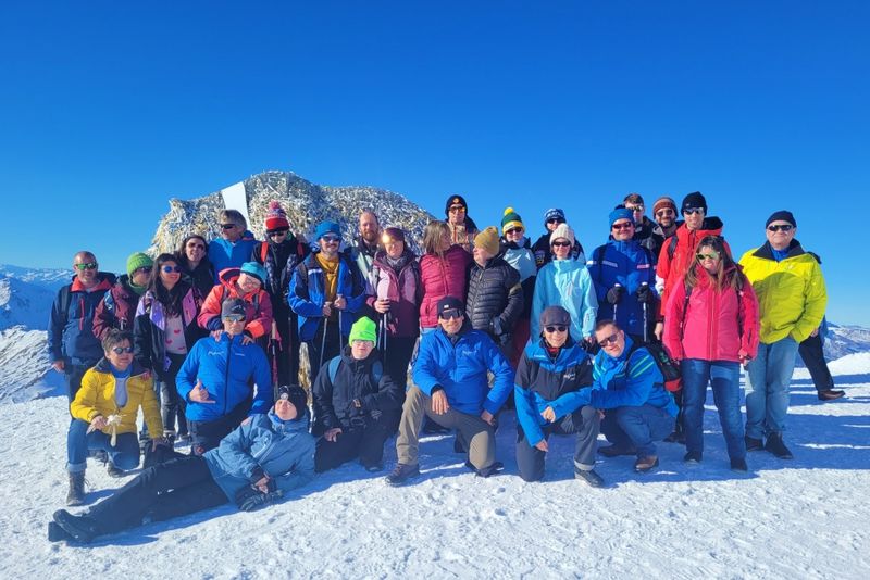 Eine Gruppe von Menschen in farbenfroher Winterkleidung posiert gemeinsam auf einem verschneiten Berg mit klarem, blauem Himmel und einem felsigen, schneebedeckten Gipfel im Hintergrund.