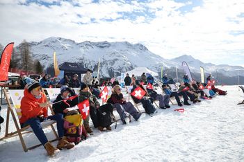 Menschen sitzen auf Liegestühlen im Schnee und schwenken Schweizer Fahnen. Im Hintergrund sind schneebedeckte Berge und ein teilweise bewölkter Himmel zu sehen, mit Zelten und anderen Zuschauern.