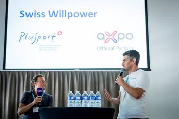 Zwei Männer sprechen auf der Bühne einer Veranstaltung, der eine hält ein Mikrofon und steht, der andere sitzt mit einem Mikrofon. Hinter ihnen ist ein Bildschirm mit Swiss Willpower und den Logos von PluSport und Axpo Official Partner zu sehen.