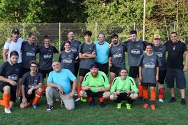 Eine Fußballmannschaft stellt sich für ein Gruppenfoto auf einer Wiese auf. Die Spieler in grauen Trikots und orangefarbenen Stutzen stehen und knien neben Trainern und Fans, im Hintergrund sind Bäume und ein Maschendrahtzaun zu sehen.