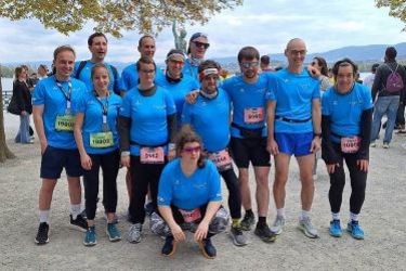 Eine Gruppe von zwölf Personen in blauen Hemden und Startnummern posiert gemeinsam im Freien bei einer Laufveranstaltung, mit Bäumen, Wasser und anderen Teilnehmern im Hintergrund.
