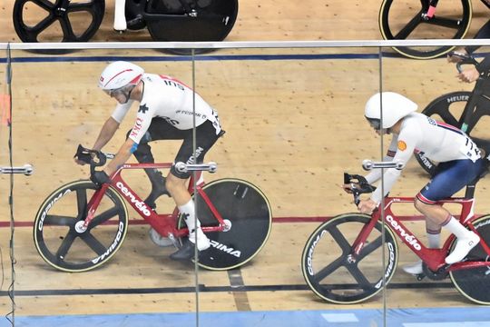 Zwei Radfahrer in Helmen und Rennanzügen fahren mit roten Rennrädern auf einem Velodrom, von oben durch eine Glaswand betrachtet, wobei im Hintergrund eine hölzerne Bahn und Fahrradräder zu sehen sind.