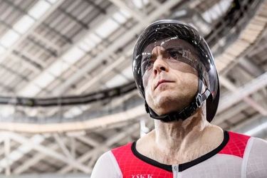 Ein männlicher Bahnradfahrer in rot-weißer Schweizer Uniform und Helm steht unter dem hellen Dach einer Velodromhalle und sieht konzentriert und rennbereit aus.