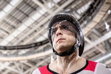 Ein männlicher Bahnradfahrer in rot-weißer Schweizer Uniform und Helm steht unter dem hellen Dach einer Velodromhalle und sieht konzentriert und rennbereit aus.