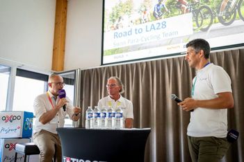 Drei Männer sitzen und stehen auf einer Bühne unter einer Leinwand mit der Aufschrift Road to LA28 Swiss Para-Cycling Team. Einer spricht in ein Mikrofon, während die anderen zuhören. Mehrere Wasserflaschen und Veranstaltungsbanner sind zu sehen.