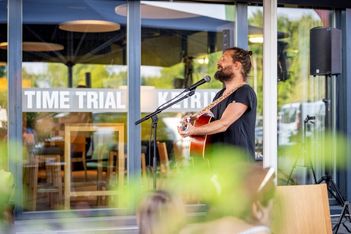 Ein Mann mit Bart und zurückgebundenem Haar singt und spielt eine akustische Gitarre in ein Mikrofon vor einem modernen Café mit großen Fenstern. Im Vordergrund ist unscharfes Grün zu erkennen.