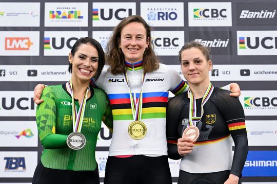 Drei Radsportlerinnen stehen auf einem Podium, lächeln und tragen Medaillen. Die Athletin in der Mitte trägt ein weißes Trikot mit Regenbogenstreifen und eine Goldmedaille. Die linke Athletin trägt grün, die rechte schwarz; beide halten Silber- und Bronzemedaillen.
