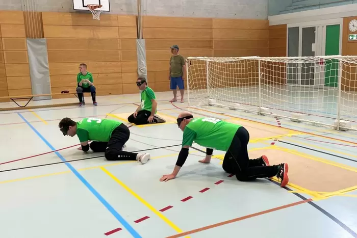 Drei Sportler in grünen Hemden und schwarzen Hosen mit verbundenen Augen knien und kriechen auf dem Boden einer Turnhalle in der Nähe eines Tornetzes. Ein Trainer steht in der Nähe und schaut zu. Die Szene spielt sich in einer Sporthalle ab.