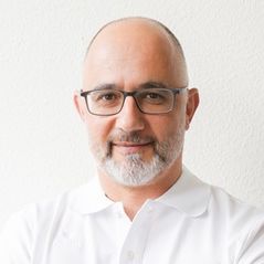 Ein Mann mittleren Alters mit Glatze, grauem Bart und Brille, der ein weißes Hemd mit Kragen trägt, steht vor einer schlichten weißen Wand und lächelt sanft in die Kamera.