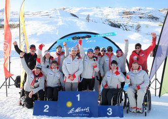 Eine Gruppe von Athleten und Trainern in Wintersportkleidung, darunter zwei Rollstuhlfahrer, feiern auf einem Siegerpodest im verschneiten St. Moritz mit Bergen und Fahnen im Hintergrund.