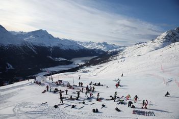 Skifahrer bereiten sich vor und ruhen sich auf einer verschneiten Piste aus, auf der die Skiausrüstung verstreut ist. Im Hintergrund sind schneebedeckte Berge und ein See bei teilweise bewölktem Himmel zu sehen.
