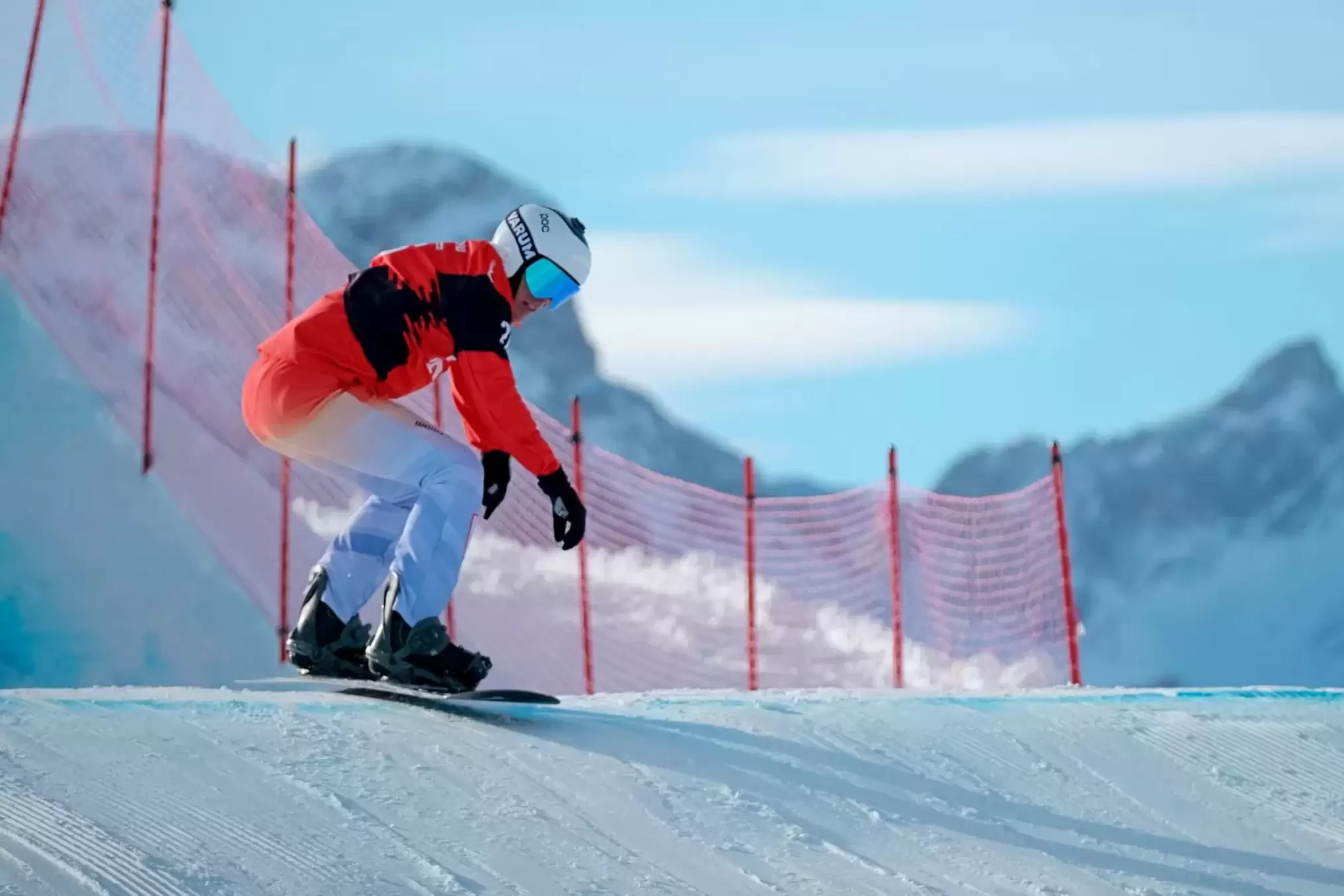 Ein Snowboarder in einem rot-weißen Anzug und einer blauen Brille fährt eine verschneite Piste hinunter, mit einem roten Netzzaun und schneebedeckten Bergen im Hintergrund unter einem klaren Himmel.