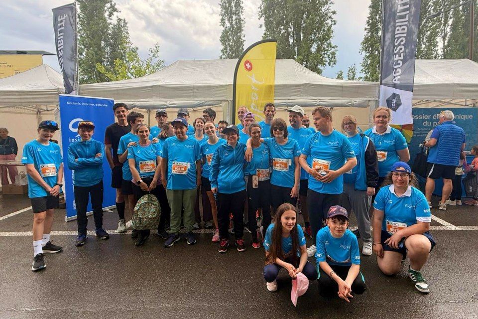 Eine Gruppe von Menschen in passenden blauen Shirts und Startnummern posiert gemeinsam im Freien bei einer Laufveranstaltung. Im Hintergrund sind Zelte und Banner zu sehen. Einige Personen stehen, andere knien oder sitzen davor.