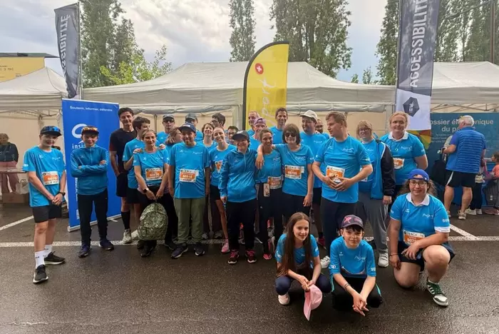 Eine Gruppe von Menschen in passenden blauen Shirts und Startnummern posiert gemeinsam im Freien bei einer Laufveranstaltung. Im Hintergrund sind Zelte und Banner zu sehen. Einige Personen stehen, andere knien oder sitzen davor.