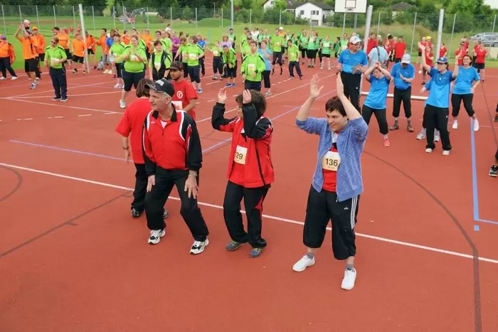Eine Gruppe von Menschen in bunten Sportuniformen trainiert gemeinsam auf einem Platz im Freien. Sie stehen in Reihen, heben ihre Arme und bewegen sich synchron, was auf eine Gruppenfitness oder Aufwärmübung hindeutet.