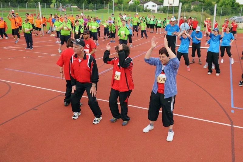 Eine Gruppe von Menschen in bunten Sportuniformen trainiert gemeinsam auf einem Platz im Freien. Sie stehen in Reihen, heben ihre Arme und bewegen sich synchron, was auf eine Gruppenfitness oder Aufwärmübung hindeutet.