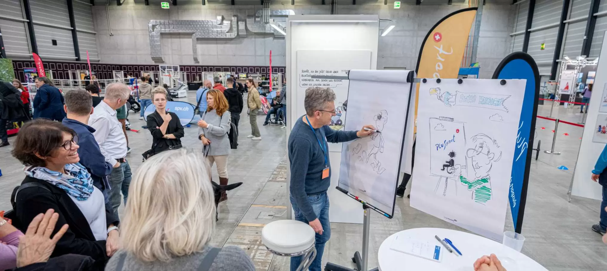 Eine Gruppe von Menschen beobachtet einen Mann, der bei einer Indoor-Veranstaltung Cartoons auf großen Flipcharts zeichnet. Die Szene spielt sich in einer geräumigen Halle mit verschiedenen Ständen und Bannern im Hintergrund ab.