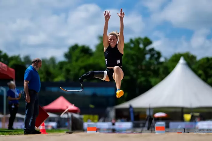 Eine Sportlerin mit einer Beinprothese befindet sich während eines Weitsprungs bei einer Leichtathletikveranstaltung im Freien mit erhobenen Armen in der Luft, im Hintergrund sind Zuschauer und Zelte unter einem teilweise bewölkten Himmel zu sehen.