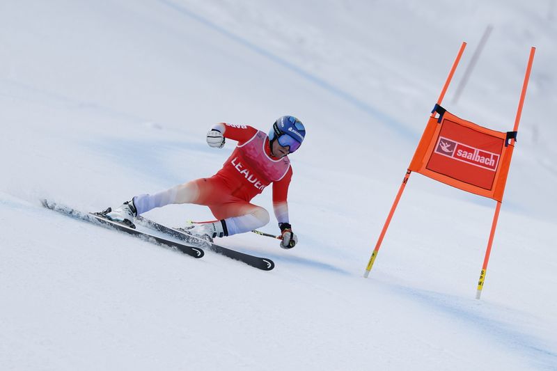 Ein Skifahrer in einem roten Anzug und mit blauem Helm macht eine scharfe Kurve um ein orangefarbenes Tor mit der Aufschrift Saalbach auf einer verschneiten Abfahrtsstrecke.