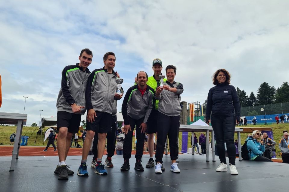 Sechs Personen, fünf eng beieinander stehende Personen in Sportkleidung und eine einzeln stehende Person, posieren auf einer Bühne im Freien bei einer Sportveranstaltung. Der Hintergrund zeigt eine Laufbahn, einen grasbewachsenen Hügel, Zelte und Menschen unter einem bewölkten Himmel.