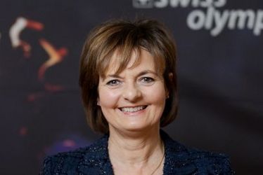 Eine Frau in einem dunklen Blazer lächelt vor einer Kulisse von Swiss Olympic, die eine Fackel und das Logo von Swiss Olympic zeigt.