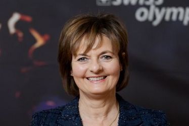 Eine Frau in einem dunklen Blazer lächelt vor einer Kulisse von Swiss Olympic, die eine Fackel und das Logo von Swiss Olympic zeigt.
