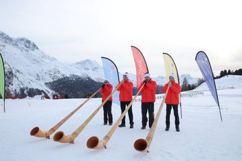 Vier Personen in roten Jacken spielen auf langen Alphörnern im Schnee, mit bunten Fahnen hinter ihnen. Im Hintergrund sind verschneite Berge und Kiefern unter einem bewölkten Himmel zu sehen.