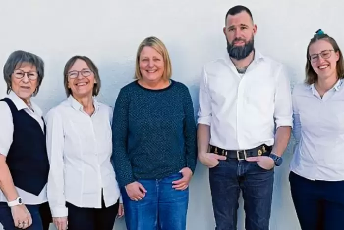 Fünf Erwachsene stehen in einer Reihe vor einem schlichten weißen Hintergrund und lächeln. Vier Frauen tragen legere Oberteile und Brillen, während ein Mann mit Bart und Tattoos ein weißes Hemd und Jeans trägt, die Hände in den Taschen.