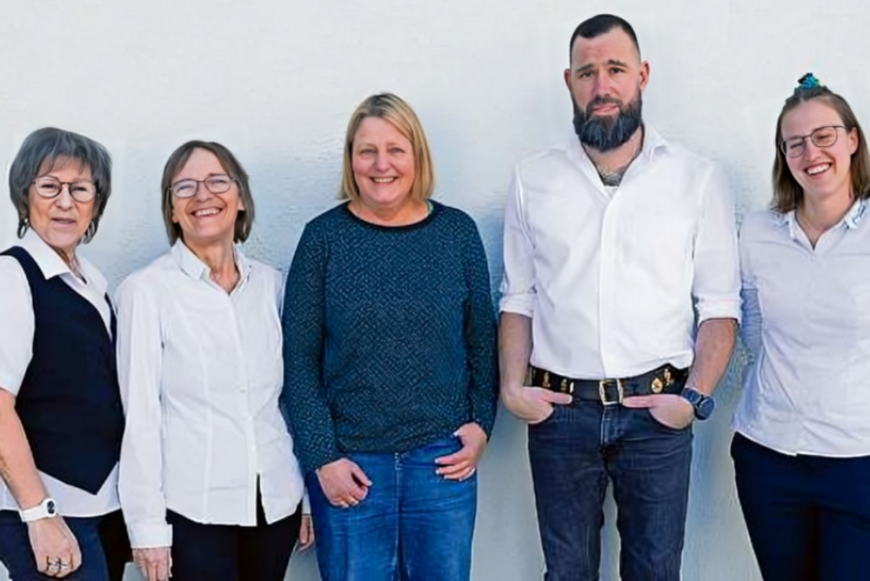 Fünf Erwachsene stehen in einer Reihe vor einem schlichten weißen Hintergrund und lächeln. Vier Frauen tragen legere Oberteile und Brillen, während ein Mann mit Bart und Tattoos ein weißes Hemd und Jeans trägt, die Hände in den Taschen.